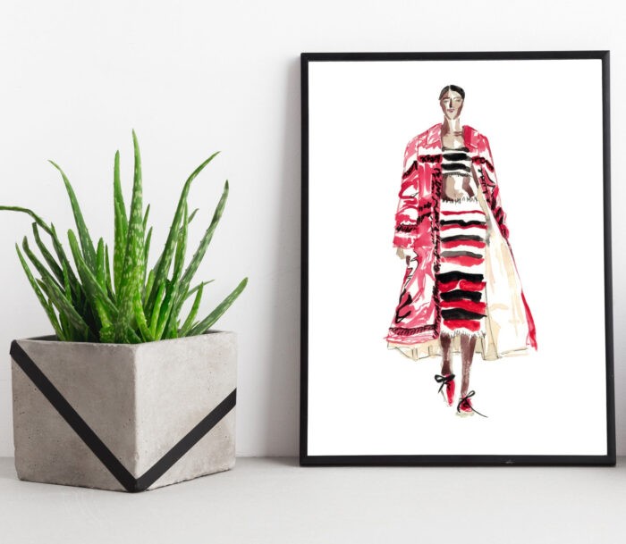A framed fashion drawing leans on a table beside a plant