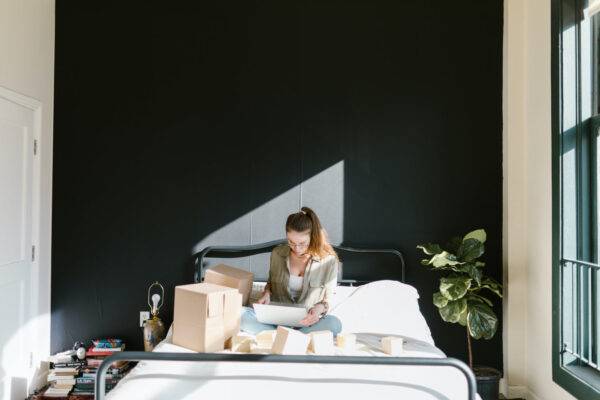 Woman sitting on a bed surrounded by boxes, working on a laptop