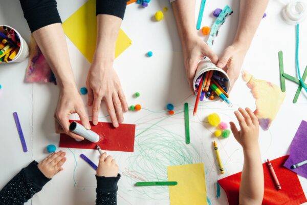 Top view of a table where a bunch of hands are playing with messy colorful crafts