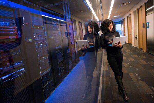 woman in carpeted hallway holding laptop while leaning against glass window of server room