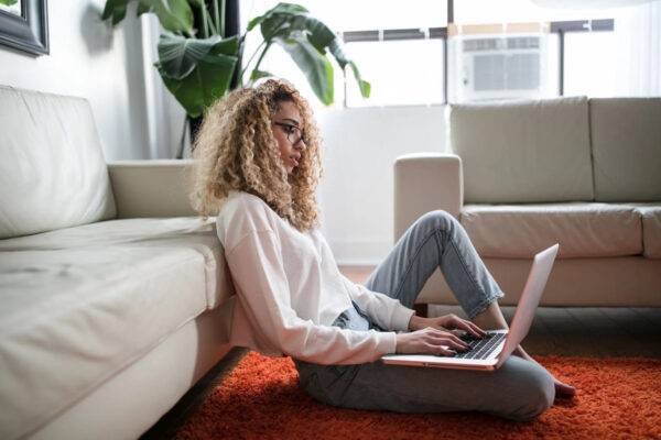 Woman with curly hair using laptop on carpeted floor next to couch and plant