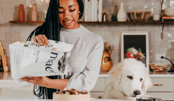 A fluffy dog waits while their owner pours Maev raw food formula into a bowl from a large bag.