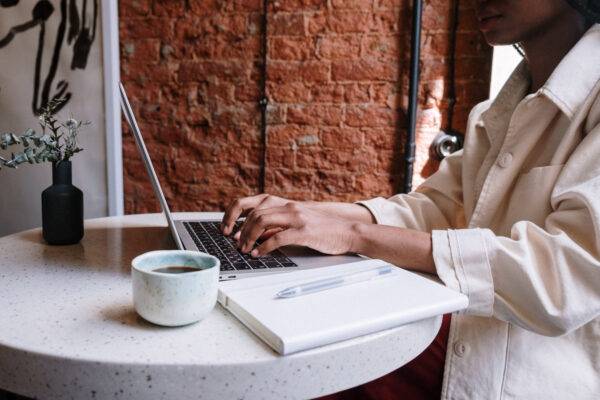 Person typing on a laptop at a table with a notebook and coffee cup, against a brick wall.