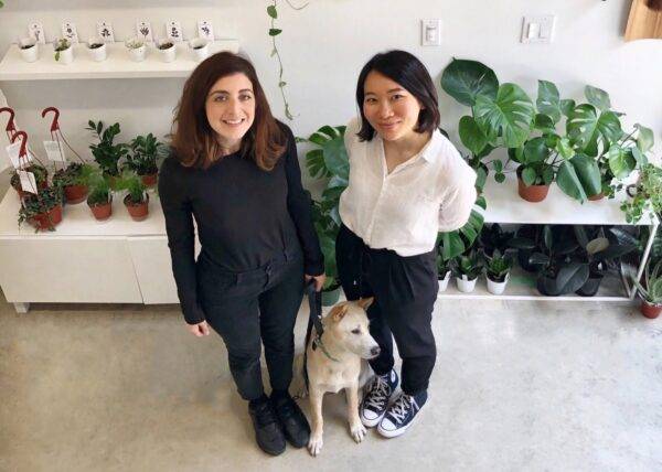 Two women and a small dog stand in front of rows of plants on white shelving