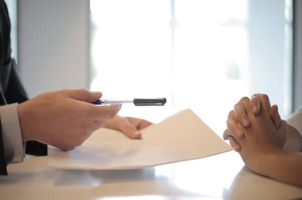 Hands holding paper and pen across from folded hands at a desk.